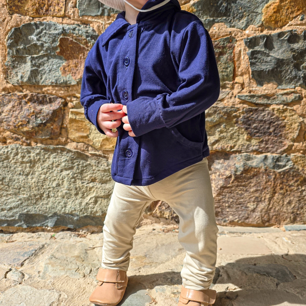 A dark navy blue Sun Baby button-up shirt, with large pockets on each side of the buttons a the bottom of the shirt and a high collar neck along with extra hand coverage at the end of the sleeves with a slit for the thumb, hanging on a clothesline against a background of a blue sky with white clouds.