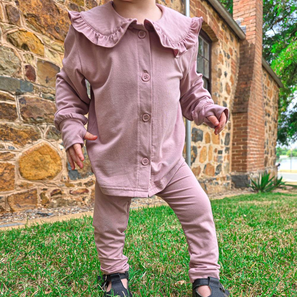 A dusky pink Sun Baby button-up shirt, with a frilly high collar neck along with extra hand coverage at the end of the sleeves with a slit for the thumb, hanging on a clothesline against a background of a blue sky with white clouds.
